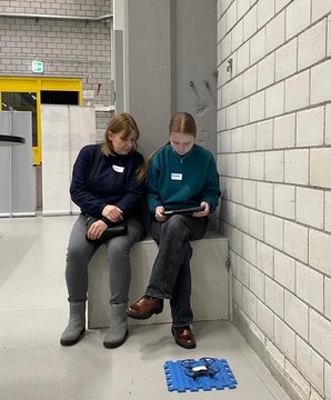 Zwei Personen sitzen in einem Raum mit Ziegelwänden und betrachten ein Tablet.